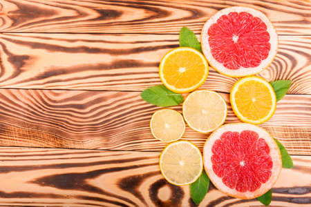 Two juicy, fresh and bright red grapefruit slices and two slices of ripe, sweet and delicious oranges and three small, round, appetite and sour lobes of lemon with green leaves on a light wooden tableの写真素材