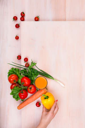 Multi-colored vegetables on a light brown wooden table. A woman hand holding a fresh yellow salad pepper. Several ripe tomatoes, carrot, onions, parsley, lettuce, salad peppers on the table.の写真素材