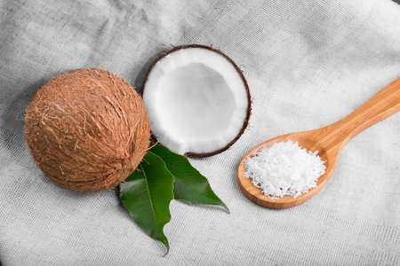 Fresh whole and crushed coconuts, light brown wooden spoon with juicy nut chips and bright green leaves on a gray fabric. Tropical coconuts with leaves. Coconut chips or flakes.の写真素材