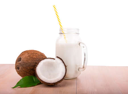 A huge mason jar with handle full of coconut milk and two tropical coconuts with leaf on a light wooden table, isolated on a white background. Whole and cut hawaiian coconuts. Fresh coconut milk.の写真素材
