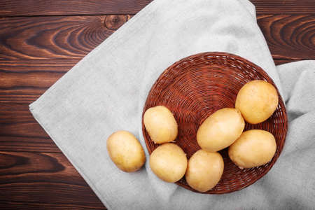 Peeled potato tubers on a bright wooden background. A brown basket full of delicious fresh potatoes on a silvery burlap sack. Uncooked raw vegetables full of nutrients.の写真素材