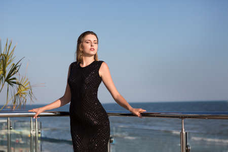 A stunning confident woman posing on a glass terrace on a bright blue natural background. An elegant female in a saturated black dress on an amazing trip. A beautiful view on a sunny sea.の写真素材