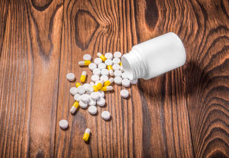 Pharmaceutical medicine pills, tablets, and capsules on a dark background. Open prescription bottle with white and yellow vitamins and drugs scattered on the table.の写真素材