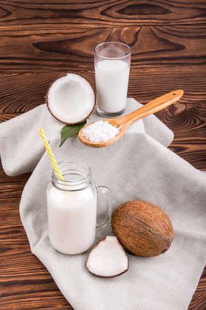Mason jar and a glass full of fresh coconut milk with green leaves and wooden spoon with coconut chips on a light fabric and on a dark wooden background. A coco, coconut milk and nut chips.の写真素材