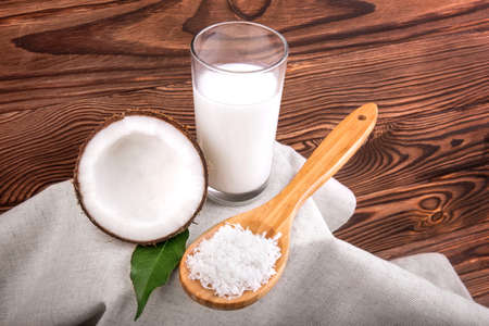 Exotic coconuts. A large glass of tasty coco milk and wooden spoon with grated nuts and half fresh tropical coconut with a bright green leaf on a light gray cloth and a dark brown wooden table.の写真素材