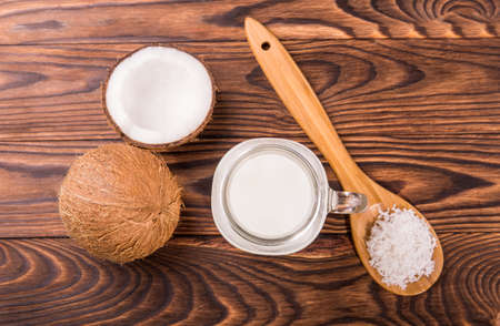 Tasteful tropical coconuts with a glass full of organic fresh milk and wooden spoon with coconut chips on a dark wooden background. Nutritious cracked coconuts on a brown table. Summer fruits.の写真素材