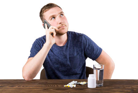 Serious young man in a dark blue T-shirt talking on the phone, blisters with tablets and a glass of water on the table, isolated on a white background. The man with pills or medicine are on the brown table.の写真素材