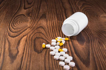 An open container with white and yellow pills or antibiotics on a wooden table. Pills, tablets, vitamins, medicine, and drugs on the desk. Healthful and medicinal drugs.の写真素材