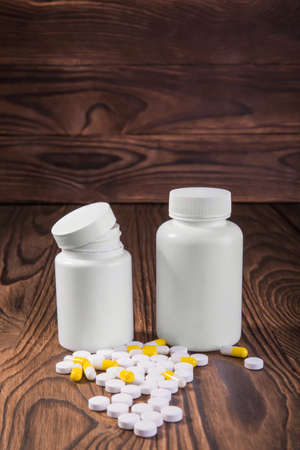 Close-up picture of two big white medicine bottles with painkillers, vitamins, and pills on a brown wooden background. A small pile of different color tablets near the bottles of medicines.の写真素材