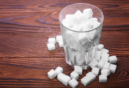 A glass of sugar crystals or cubes on a dark brown wooden table. A few sugar cubes are near the full glass with white sugar. Intake of bad calories.の写真素材