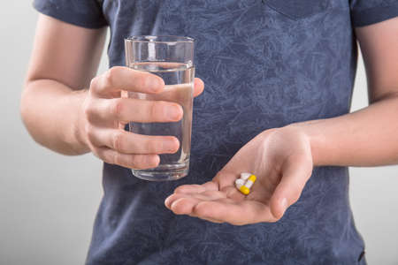 A young man in a dark blue T-shirt is holding a glass of water and few tablets of different colors on a gray background. The man drinks tablets. Suicide. Medicinal tablets.の写真素材