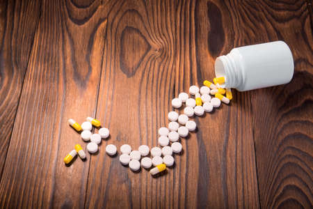 White and yellow different pills, tablets, vitamins, and drugs scattered near an opened white medicine bottle on a wooden background. Treatment with pills and medicine.の写真素材