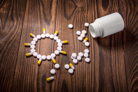 An open medical bottle and a pile of pills for treatment on a brown wooden background. A composition of capsules and tablets in the form of the sun on the desk. Treatment with pills and medicine.の写真素材