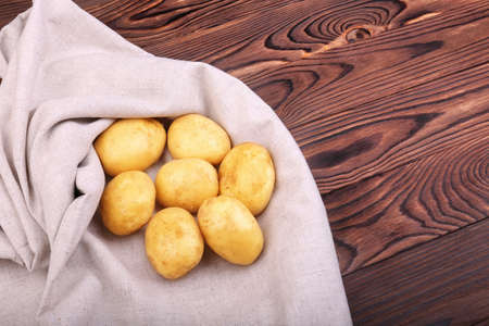 Uncooked potatoes on a dark brown wooden background. Potato tubers in a brown basket on a gray piece of cloth. Healthful rustic vegetables. Summer harvest. Peeled potatoes.の写真素材
