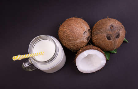 Top view of ripe, fresh, appetizing whole and cut coconuts and mason jar with coconut milk on a dark purple background. A straight and long yellow straw in the transparent mason jar.の写真素材