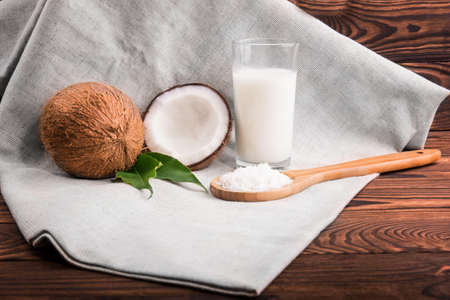 A transparent glass of coconuts, delicious coco milk, wooden spoon with coco chips on a gray bag on a wooden table. Whole and half fresh tropical coconuts with green leaves on the desk.の写真素材