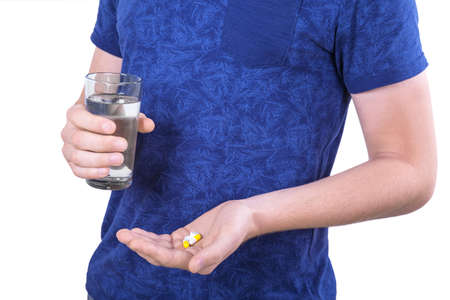 A man standing and holding medicine isolated over the white background. A male taking pills, drugs and holding a glass of clear water. A guy in a saturated dark blue t-shirt.の写真素材