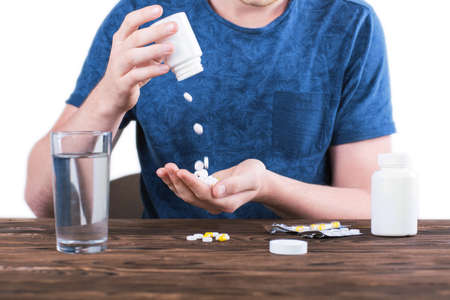 Stressed young man in a dark blue t-shirt spilling pills in his hands isolated over the white background. Bottle of tablets and a glass of clear water standing on a wooden table.の写真素材
