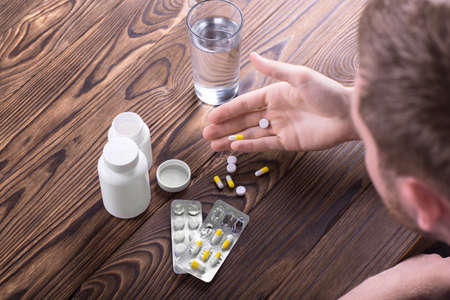 A top view of a young man looking at pills in his hand. Various drugs and painkillers in packs and a white bottle on a light brown table. A sick patient taking prescripted drugs.の写真素材