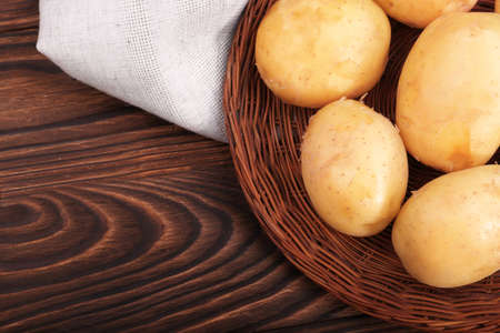 Close-up multiple raw peeled potatoes in a brown basket on a gray sack. Delicious potato tubers on a dark wooden background. A top view on uncooked bright healthful potatoes.の写真素材