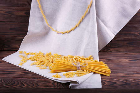 A beautiful composition of uncooked and dry pasta on a gray fabric and on a dark brown wooden table. A variety of types and shapes of dry Italian pasta on the desk.の写真素材