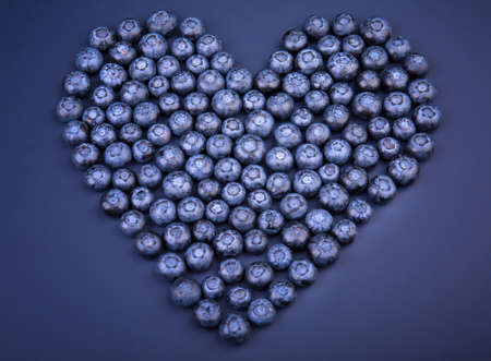 A top view of a group juicy blueberries organized in a shape of a heart on a saturated blue background. Sweet blueberries for healthful summer breakfasts. Beautiful vegan berries in a heart shape.の写真素材