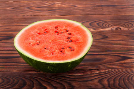 Close-up of a cut round ripe watermelon on the dark brown wooden background. The big red watermelon with a green peel and with tiny seeds. The big berry is full of vitamins.の写真素材