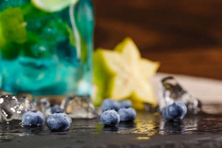 Close-up picture of a blue summer beverage on wooden background. Refreshing bright blue lagoon cocktail with mint, bilberries, ice and carambola in transparent glass. Summer beverages.の写真素材