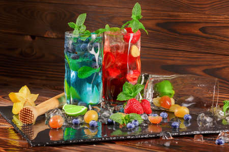 Beautiful blue and red beverages on a black tray. Bright fresh cocktails with mint, lime segments, berries, ice and carambola in transparent highball glasses on the wooden background.の写真素材