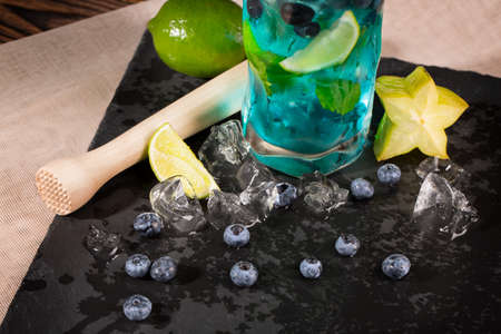 Close-up of blue fresh beverage on a black table-napkin and wooden table. Refreshing bright blue lagoon cocktail with slices of lime, bilberries, ice and carambola in transparent highball glass.の写真素材