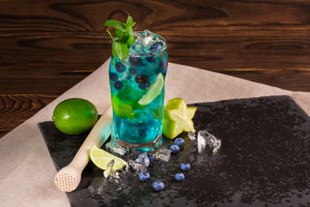 Close-up of blue fresh beverage on a black table-napkin and wooden background. Cool bright blue lagoon cocktail with lime segments, mint, bilberries, ice and carambola in huge glass. Summer beverages.の写真素材