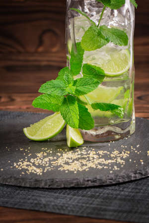 Close-up of a tasty organic beverage on a black desk with fresh lime segments, ice cubes and mint twigs on a wooden background. Beverages for summer parties. Alcoholic cocktails. Copy space.の写真素材