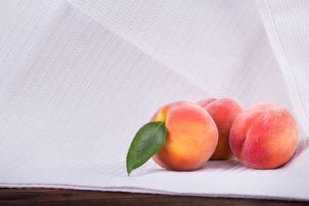 Close-up of a three whole and juicy peaches on a white table-cloth. Appetizing fruits of peaches or nectarines with green leaves full of vitamins on a light fabric. Summer fruits. Vegetarian lifestyleの写真素材