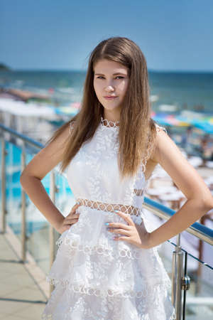A charming woman is posing on a transparent balcony on a blue sky background. The amazing and perfect girl on a summer vacation. The cute female with long light brown hair in a white dress.の写真素材