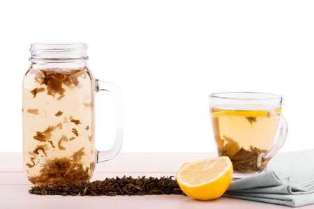 A sweet black tea and a cut lemon isolated on a white background. Organic Chinese tea in a big mason jar and a transparent cup with a hot drink. Herbal tea leaves and a lemon on a fabric on a table.の写真素材
