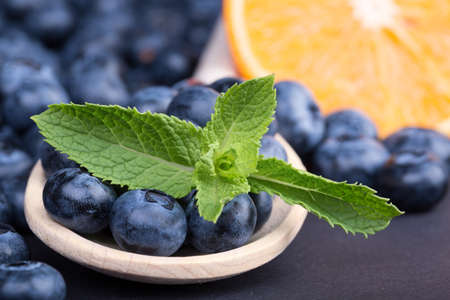 A close-up picture of juicy blueberries and mint leaves on a black background. Sweet and healthy blueberries in a little wooden spoon and a cut orange. Summer berries for a nutritious breakfast.の写真素材