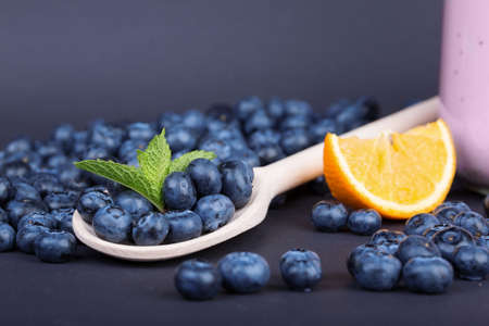 A close-up picture of juicy blueberries and mint leaves on a black background. Sweet and healthy blueberries in a little wooden spoon and a cut orange. Summer berries for a nutritious breakfast.の写真素材
