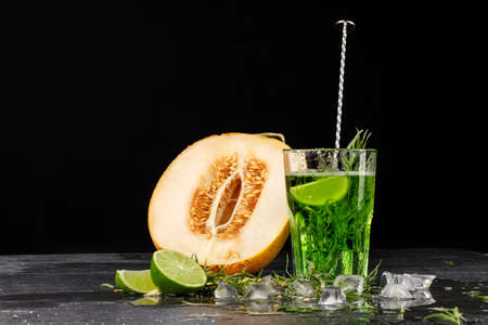 A green drink with lime and tarragon. A cocktail and a melon on a black background. Healthy, sweet and tasty drinks. Copy space.の写真素材
