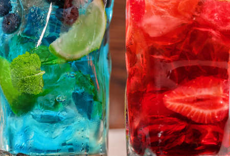 Close-up picture of blue and red drinks. Two alcohol summer cocktails with mint leaves, cut strawberries and ice in highball transparent glasses on the wooden background. Cocktails for parties.の写真素材