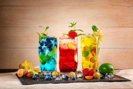 Close-up picture of blue, yellow and red drinks on black desk. Tasty bright summer cocktails with mint, blueberries, strawberries, grapes, ice and carambola in highball glasses on a wooden background.の写真素材