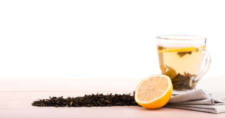 A close-up of a glass tea cup full of green tea isolated on a white background. A cup with liquid, cut lemon and green tea leaves on a table-napkin and light wooden table. Healthy beverage. Copy spaceの写真素材