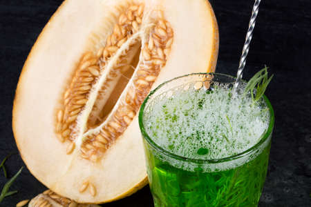 Close-up of a beautiful composition of a glass with alcoholic drink and delicious cut melon on a saturated black background. Refreshing and fruity summer beverage with ice and tarragon leaves.の写真素材