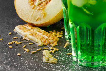 Closeup of glasses of alcoholic drink with ice and tarragon leaves and delicious cut melon on a black background. Refreshing and fruity non-alcoholic summer beverages. Fresh melon and cold cocktails.の写真素材