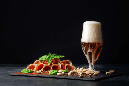 A delicious Italian ham cured by drying and served in thin slices on the saturated black background. A glass of intoxicating beer with foam. Tasty prosciutto and pistachios on the table.の写真素材
