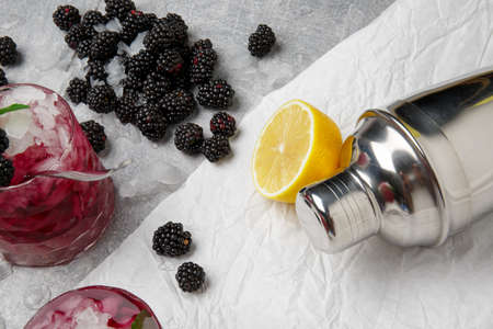 A view from above on composition of two glasses of fruit non-alcohol beverages with ice and blackberries on a gray background. A metallic shaker, berries and sliced sour lemon on the light background.の写真素材