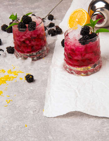 Two transparent glasses of refreshing cocktails with natural blackberries, sour yellow lemon and mint on a light background. Many berries, lemon citron, ice and shaker on the table.の写真素材