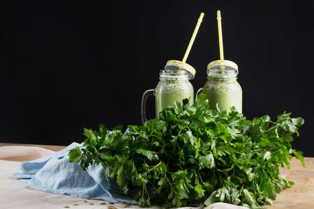 Beautiful composition of mason jars with green vegetable cocktail in a bowl with fresh parsley leaves on a wooden table on a black background. Cold, tasteful and nutritious protein shakes. Copy space.の写真素材