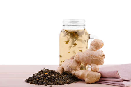 A big mason jar filled with black tea and spicy ginger on a light table isolated on a white background. A heap of dry green tea leaves and a pink fabric next to a drink. Freshness and health concept.の写真素材