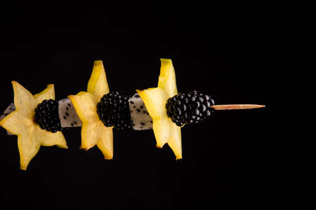 A wooden toothpick with ripe yellow carambola, dragon fruit and sweet blackberries on a saturated black background. Tasty finger food with various fruits for snack.の写真素材