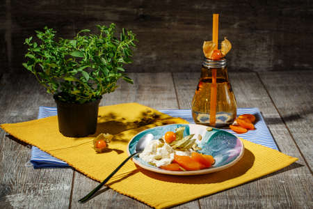 A close-up picture of a light dinner composition on colorful fabric table napkins. A blue round dish, bright orange bottle, ripe physalis, and little green bush on a wooden background.の写真素材